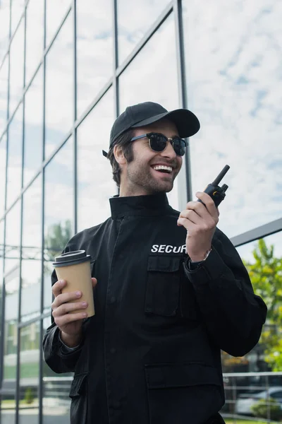 Cheerful security man with paper cup talking on walkie-talkie on city street — Stock Photo