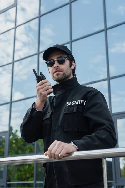 Homem de segurança em óculos de sol e uniforme preto falando no walkie talkie ao ar livre — Fotografia de Stock