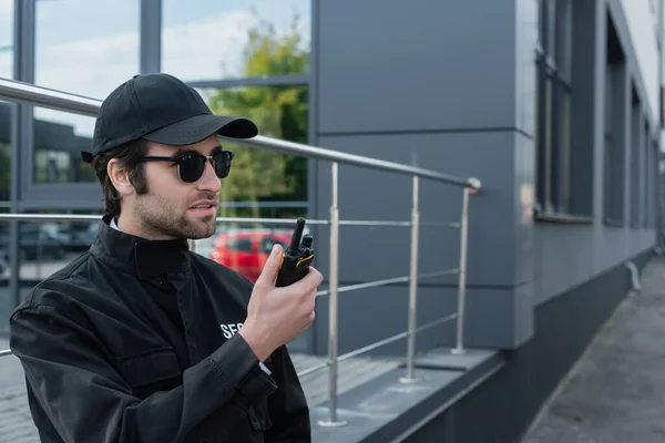 Охранник в черной форме и солнцезащитных очках разговаривает по рации возле здания — стоковое фото