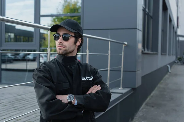 Serious guard in sunglasses and black uniform standing with crossed arms on city street — Stock Photo