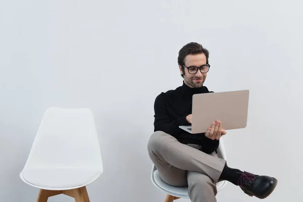 Щасливий і стильний чоловік використовує ноутбук, сидячи на стільці біля сірої стіни — стокове фото