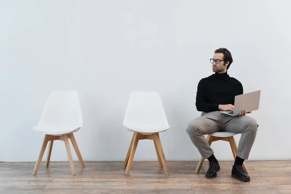 Young man in stylish clothes sitting with laptop near grey wall and looking away — Stock Photo