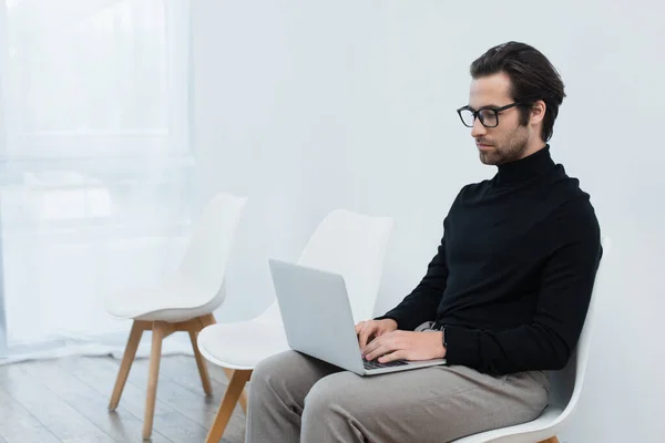 Joven freelancer en gafas y cuello alto negro sentado cerca de la pared gris y usando laptop - foto de stock