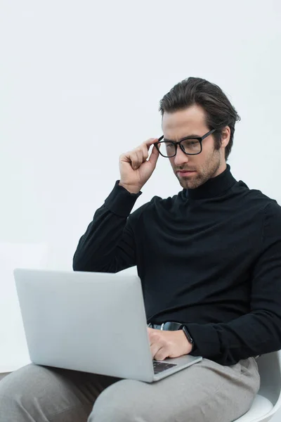 Young man in black turtleneck adjusting eyeglasses while working on laptop on grey background — Stock Photo