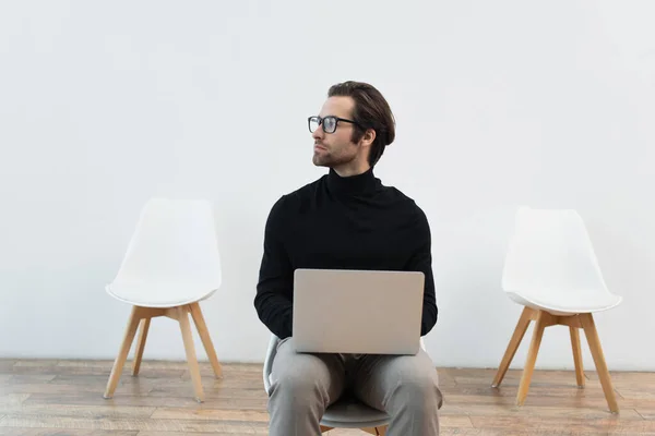 Hombre elegante sentado en la silla con el ordenador portátil y mirando hacia otro lado - foto de stock