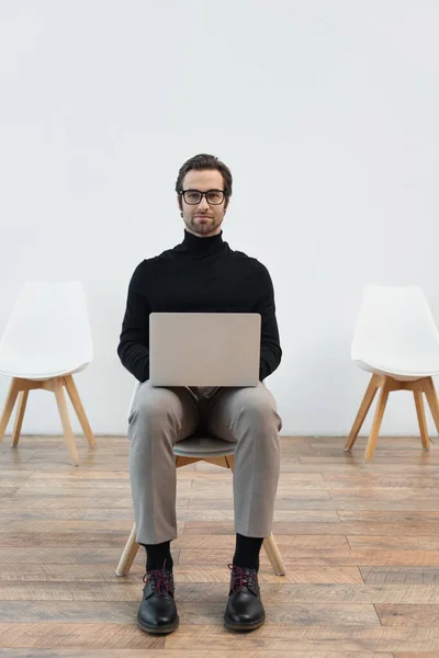 Young man in black turtleneck and eyeglasses sitting on chair with laptop and looking at camera — Stock Photo