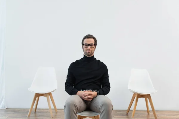 Joven y elegante hombre de cuello alto negro y gafas de vista sentado en la silla y mirando a la cámara - foto de stock