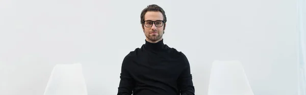 Молодий чоловік в окулярах і чорна форма дивиться на камеру біля стільців на сірому фоні, банер — стокове фото