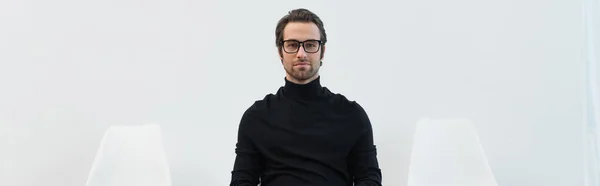 Young man in eyeglasses and black uniform looking at camera near chairs on grey background, banner — Stock Photo