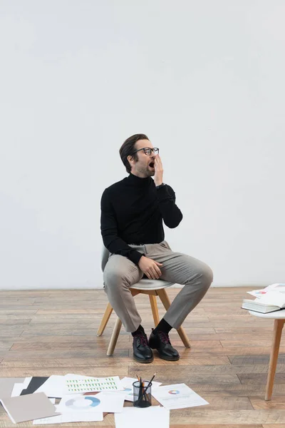 Hombre soñoliento bostezando en la silla cerca de documentos en el suelo y cuadernos en la mesa de café - foto de stock