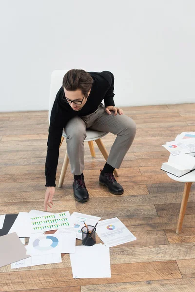 Homem sentado na cadeira e chegando a documentos com infográficos no chão — Fotografia de Stock