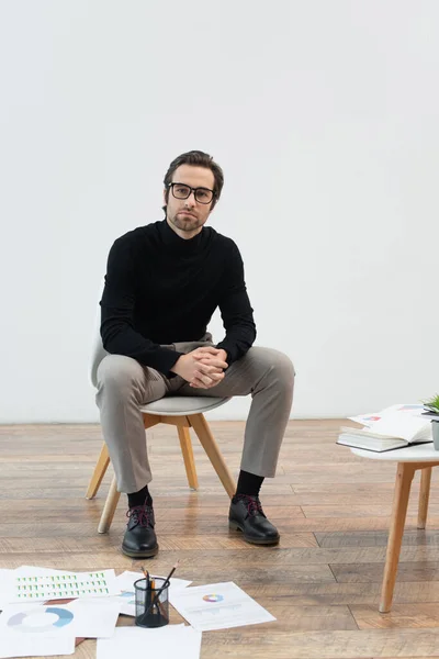 Young and stylish man sitting on chair near documents on floor and coffee table — Stock Photo