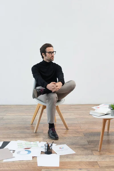 Молодий чоловік у стильному одязі, що сидить біля документів з графіками на підлозі — стокове фото