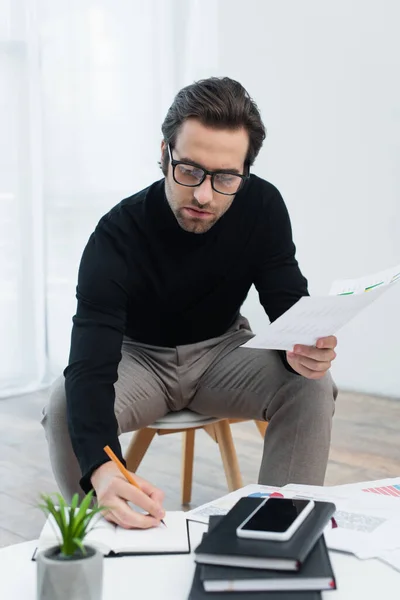 Junger Mann schreibt in Notizbuch, während er mit Papieren in der Nähe von Smartphone mit leerem Bildschirm arbeitet — Stockfoto