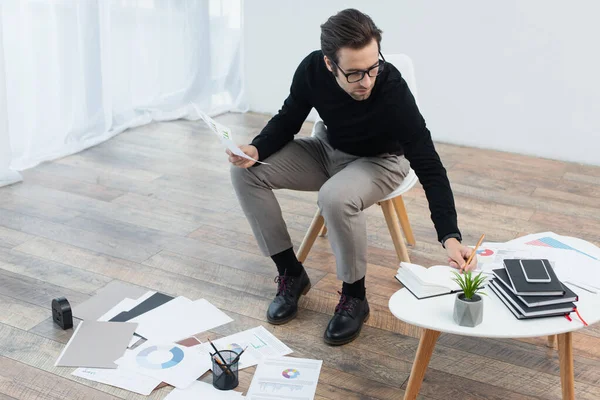 Hombre de moda escribiendo en cuaderno cerca de documentos con infografías en el suelo - foto de stock