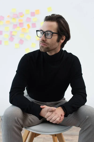 Hombre joven con cuello alto negro y anteojos sentados con las manos apretadas y mirando hacia otro lado - foto de stock