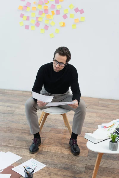 Hombre de moda mirando los documentos cerca de los papeles en el piso y la mesa de café - foto de stock