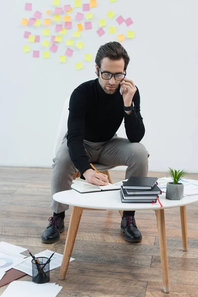Stylish man writing in notebook while talking on cellphone near papers on floor — Stock Photo
