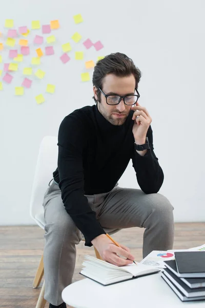 Young man talking on mobile phone and writing in notebook on coffee table — Stock Photo