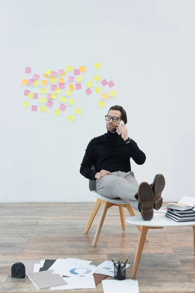 Trendy man sitting with legs on coffee table and talking on smartphone — Stock Photo