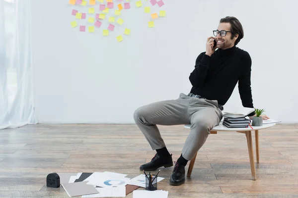 Homme heureux parlant sur téléphone portable tout en étant assis sur la table basse près des papiers sur le sol — Photo de stock