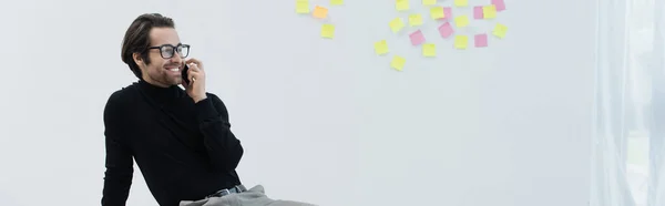 Uomo soddisfatto in occhiali che parla sul telefono cellulare vicino note appiccicose sulla parete grigia, banner — Foto stock
