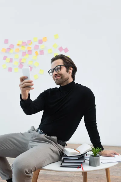 Homem feliz e elegante sentado na mesa de café perto de cadernos e tomando selfie no telefone móvel — Fotografia de Stock