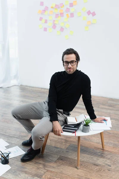 Man in black turtleneck and eyeglasses sitting on coffee table near notebooks and looking at camera — Stock Photo