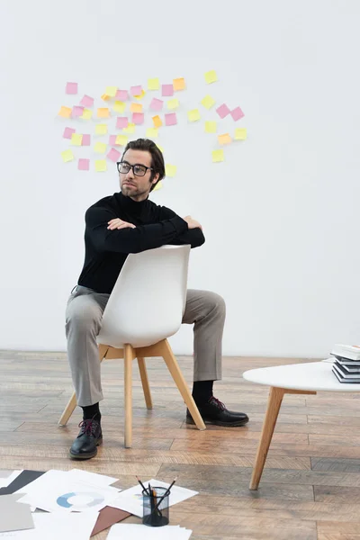 Man looking away while sitting near notebooks on coffee table and documents on floor — Stock Photo