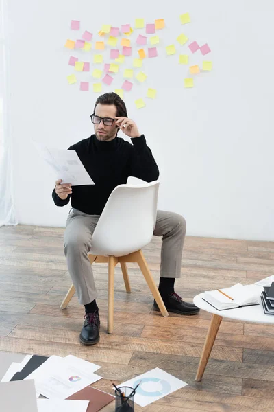 Молодий чоловік працює з документами, сидячи на стільці біля паперів на підлозі — стокове фото