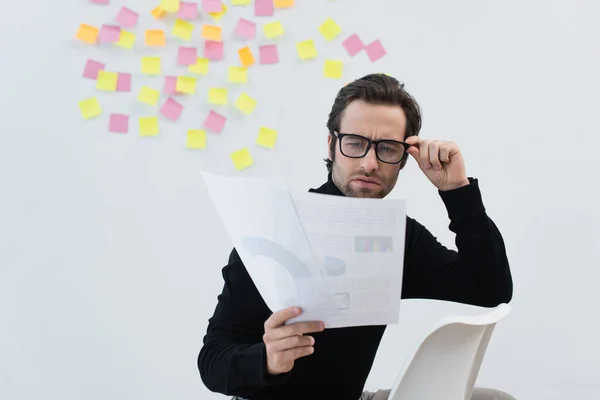 Uomo che regola gli occhiali mentre guarda i documenti vicino alla parete grigia con note adesive — Foto stock