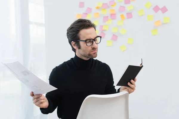 Homem em óculos e gola alta preta trabalhando com notebook e documento perto de notas pegajosas na parede cinza — Fotografia de Stock