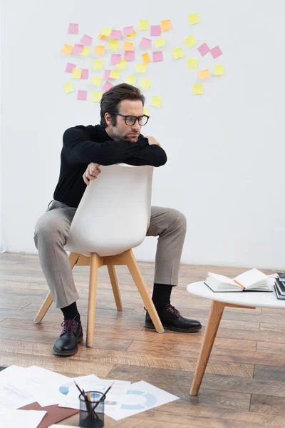 Thoughtful man sitting on chair near notebooks on coffee table and papers on floor — Stock Photo