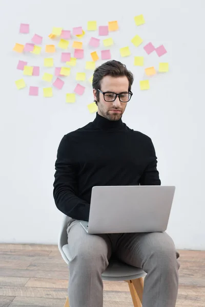Man in black turtleneck and eyeglasses working on laptop near grey wall with sticky notes — Stock Photo
