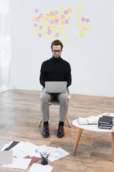 Homem da moda trabalhando no laptop perto da mesa de café com cadernos e documentos no chão — Fotografia de Stock