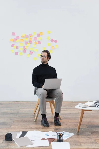 Trendy mans sitting on chair with notebook near papers on floor and notebooks on coffee table — Stock Photo