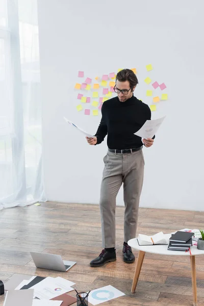 Homme élégant avec des documents debout près de l'ordinateur portable et des papiers sur le sol — Photo de stock