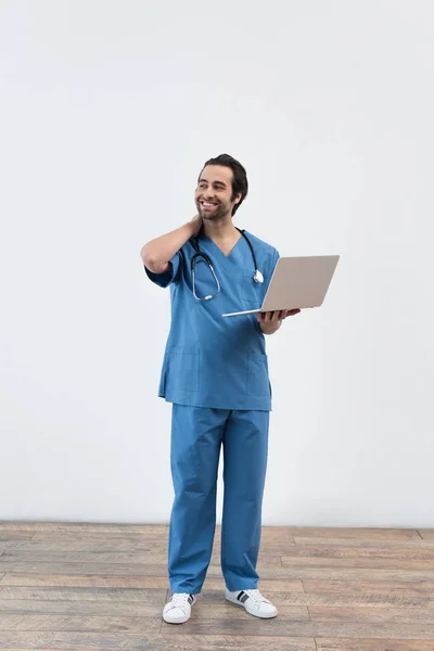 Улыбающийся врач в полный рост с ноутбуком, отводящим взгляд от серой стены — стоковое фото
