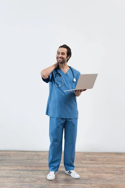 Longitud completa de médico sonriente con portátil mirando hacia otro lado cerca de la pared gris - foto de stock