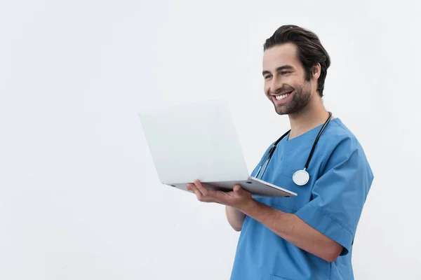 Medico soddisfatto in uniforme blu guardando computer portatile isolato su grigio — Foto stock