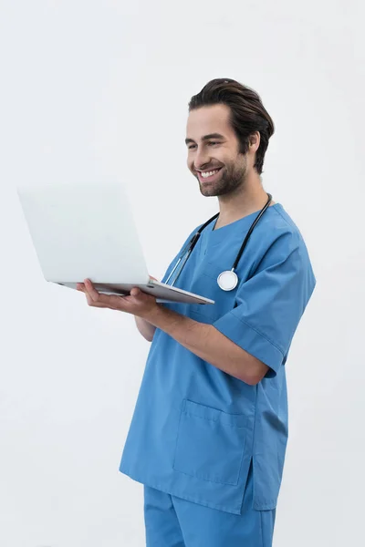 Joven médico en uniforme azul sonriendo mientras mira el ordenador portátil aislado en gris - foto de stock
