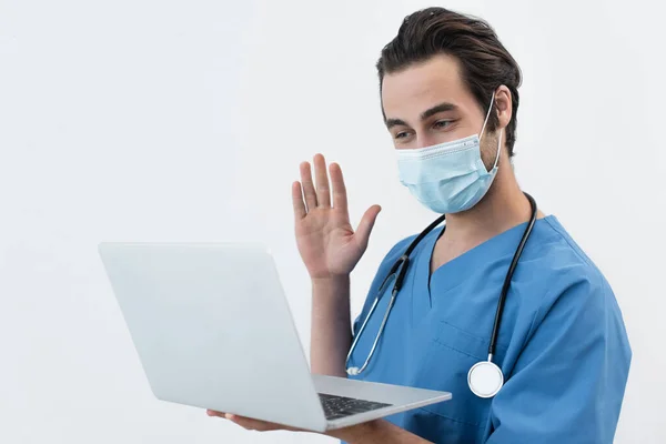 Врач в медицинской маске размахивает рукой во время видеозвонка на ноутбуке, изолированном на сером — стоковое фото