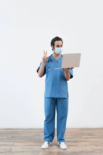 Full length view of doctor in medical mask showing victory sign during video call on laptop near grey wall — Stock Photo