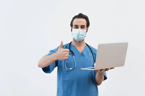 Young doctor in medical mask holding laptop and showing like gesture isolated on grey — Stock Photo