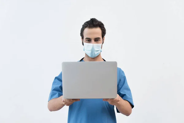 Молодой врач в медицинской маске держит ноутбук изолирован на серый — стоковое фото