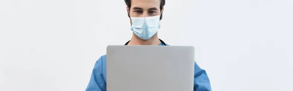 Joven médico en máscara médica utilizando portátil aislado en gris, bandera - foto de stock