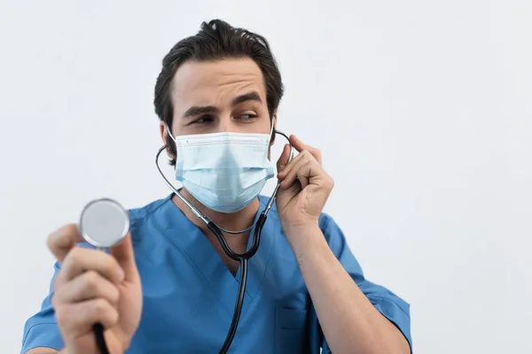 Médico reflexivo en máscara médica utilizando estetoscopio aislado en gris - foto de stock