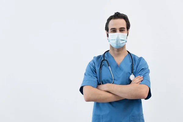 Jovem médico em máscara médica de pé com os braços cruzados e olhando para a câmera isolada em cinza — Fotografia de Stock