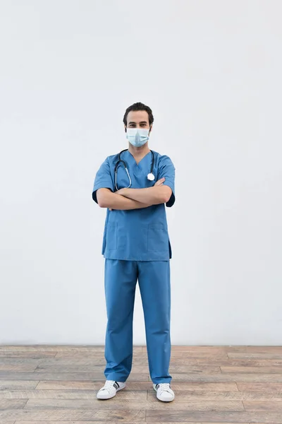 Comprimento total do médico em máscara médica de pé com os braços cruzados perto da parede cinza — Fotografia de Stock