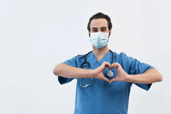 Doctor in medical mask and blue uniform showing heart sign with hands isolated on grey — Stock Photo